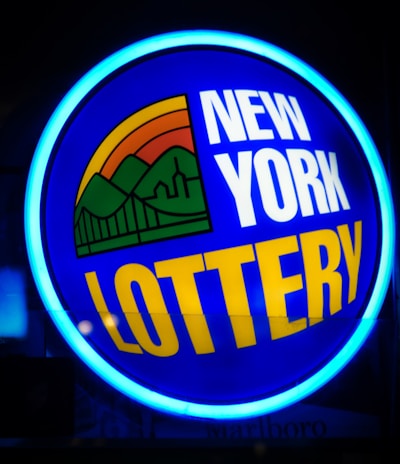 A bright, circular neon sign with a blue background, featuring the words 'New York Lottery' in bold white and yellow letters. The sign includes a colorful illustration of a landscape with green mountains, an orange-red sunset, and a bridge, all encapsulated within the circle.