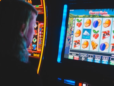 A person is closely observing a digital slot machine with colorful fruit symbols, bars, and a bell. The slot machine screen displays symbols like oranges, plums, cherries, and clouds. The background is dimly lit, creating a focused and somewhat intense atmosphere.