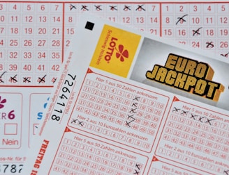 Three lottery tickets are displayed, including Eurojackpot and Super 6. Various numbers are marked on the tickets, with some crossed out and others checked. The Eurojackpot ticket is partially visible with the logo prominently displayed, and a ticket number is visible on one of the tickets.