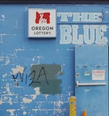 A weathered blue brick wall featuring a 'Oregon Lottery' sign with a stylized hand logo. The word 'BLUE' is painted in large letters next to a utility door with a 'No Minors Permitted' sign. There is graffiti reading 'VISA' on the wall, partially covering a green patch. An old yellow metal post and an orange traffic cone are positioned in front of the wall. A tree can be seen in the background above the wall.