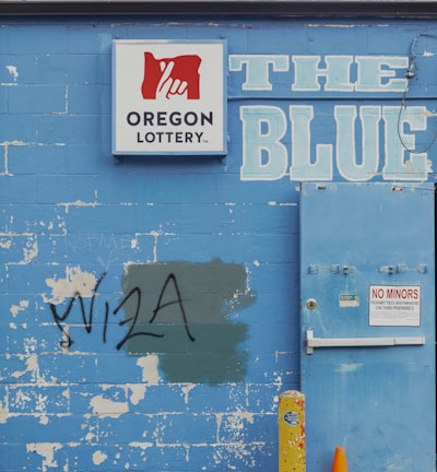 A weathered blue brick wall featuring a 'Oregon Lottery' sign with a stylized hand logo. The word 'BLUE' is painted in large letters next to a utility door with a 'No Minors Permitted' sign. There is graffiti reading 'VISA' on the wall, partially covering a green patch. An old yellow metal post and an orange traffic cone are positioned in front of the wall. A tree can be seen in the background above the wall.
