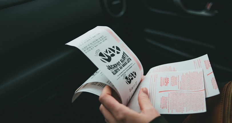 A person is holding and examining what appears to be lottery tickets inside a car. The image is focused on the tickets and the person's hands, with parts of the car interior visible in the background.