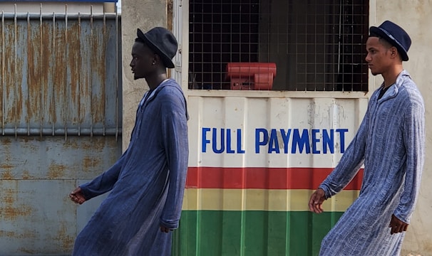 Two individuals are walking past a small kiosk with signage for the National Lottery Authority. The kiosk, decorated with a sign that reads 'Full Payment,' features yellow, green, red, and white colors. The individuals are dressed in matching long blue robes and hats, with one wearing black boots and the other white boots.