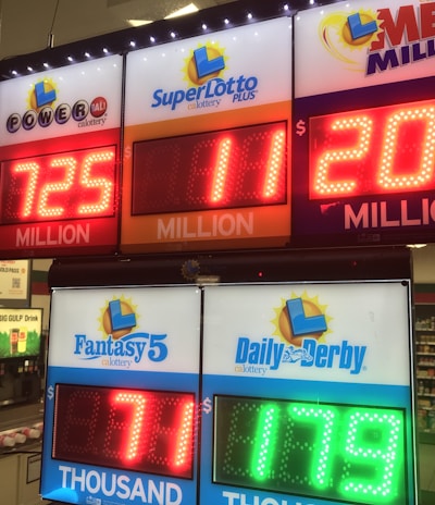 A signboard inside a convenience store displays the current jackpot amounts for various lottery games. There are bright, illuminated numbers in red and green showing the prize amounts: Powerball is at 725 million, Super Lotto Plus at 11 million, Mega Millions at 205 million, Fantasy 5 at 71 thousand, and Daily Derby at 179. In the background, shelves with various drinks and products are visible, along with promotional posters.